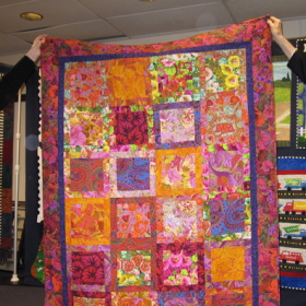 A multicolor quilt in pink, orange, yellow and purple is being displayed by two members