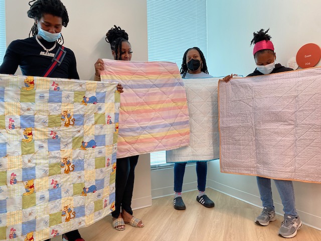 Members of DC Diaper bank hold up donated quilts from the members, including a Winnie the Pooh quilt, a quilt with pink and blue stripes, and two pink and white quilts 