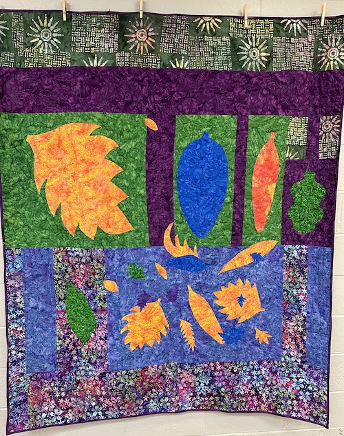 A close up of an abstract applique quilt with yellow blue and orange leaves against a background that is purple, green, and blue 