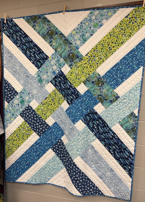 A quilt hanging on a wall with interwoven stripes of blue green and grey against a white background
