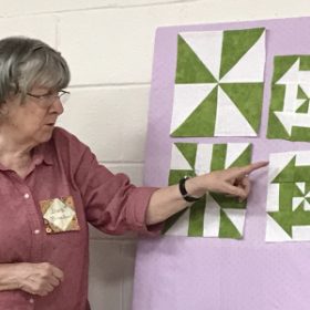 A white woman with grey hair and a plum blouse points to a display board showing four different green and white quilt blocks. She is demonstrating how the blocks were made. 