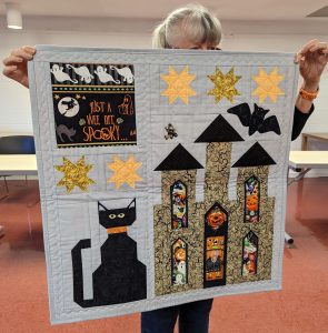 A halloween quilt with a castle, bats, a black cat, and stars. There's halloween fabric peeking out the windows of the castle, and a just a wee bit spooky fabric panel at the top left