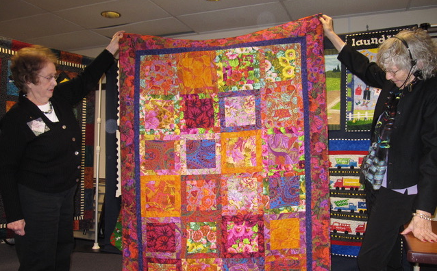 A multicolor quilt in pink, orange, yellow and purple is being displayed by two members