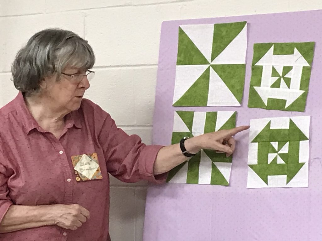 A white woman with grey hair and a plum blouse points to a display board showing four different green and white quilt blocks. She is demonstrating how the blocks were made. 